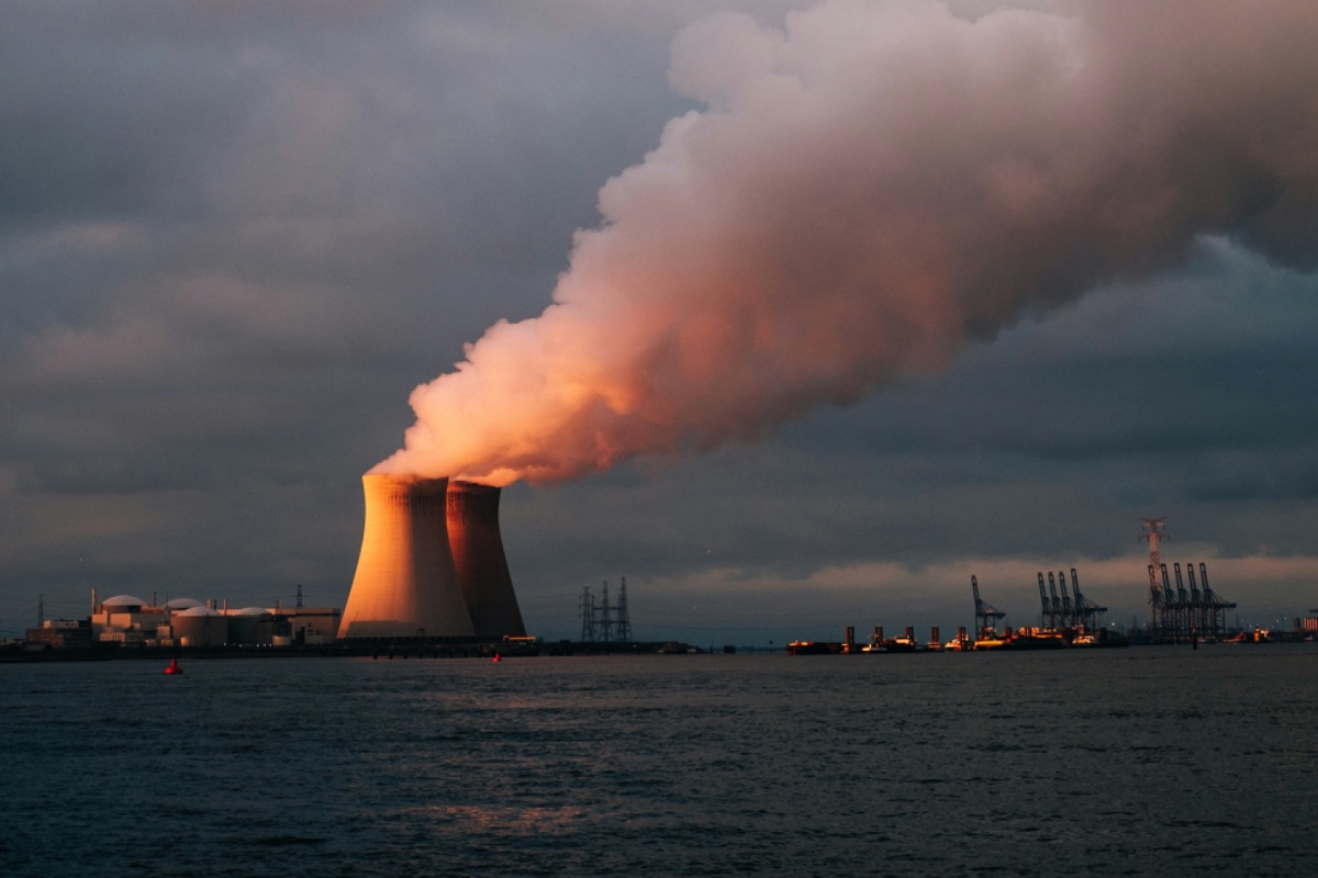 Cooling towers of a nuclear generating station releasing steam at dusk beside a working waterfront.